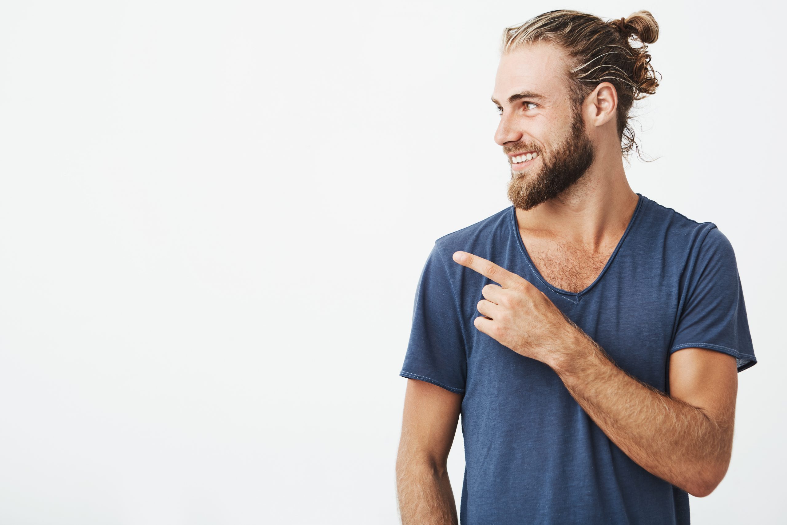 Profile of cheerful handsome man with fashionable hairstyle and beard smiling brightfully and pointing at free space for advertisement